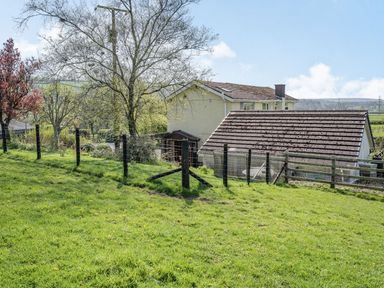A fenced grassy yard with trees and a house with a tiled roof in the background at Pant Y Cwtsh in Kidwelly