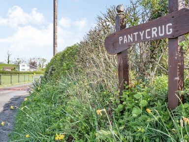 A wooden sign with pantycrug text next to bushes beside a rural road with a house and fields in the background at Pant Y Cwtsh in Kidwelly