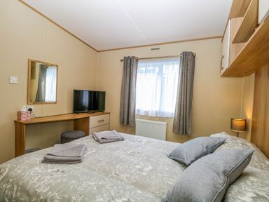 A bedroom with a bed, pillows, folded towels, a television, a mirror, a desk, a window with curtains, and a lamp at Oakley Retreat in Fritton near Belton