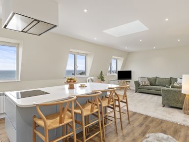 A living area with a kitchen island and wooden stools and green sofas with a TV and windows overlooking the sea at No 10 Croft House in Tenby