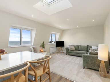 A living area with green sofas a TV on a stand by two windows and a white island with wooden chairs at No 10 Croft House in Tenby