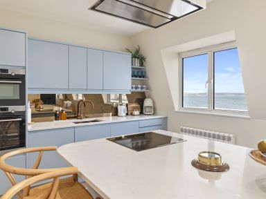 A kitchen with blue cabinets a window overlooking the sea and a breakfast bar with wooden chairs at No 10 Croft House in Tenby