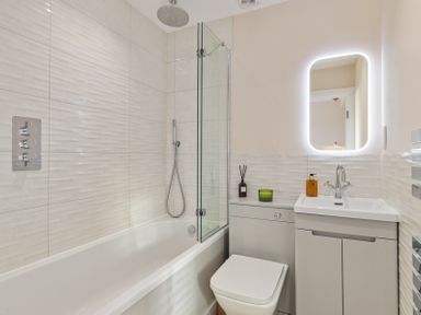 A bathroom with a bathtub shower combination a toilet a sink with cabinet and a lighted mirror at No 10 Croft House in Tenby