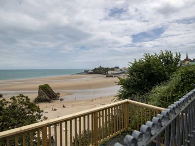 A beach with a sandy shore and rock formation viewed from a wooden balcony with trees and buildings in the distance at No 10 Croft House in Tenby