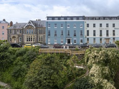 A row of buildings with parked cars in front and a green hillside with stairs and a wooden deck at No 10 Croft House in Tenby