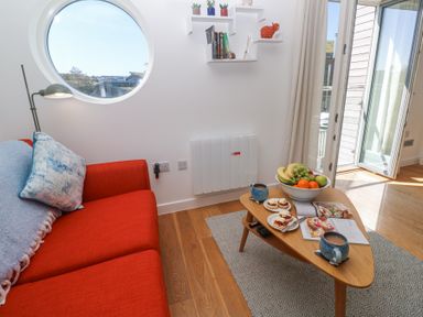 A sitting area with an orange sofa, a wooden coffee table with mugs, fruit bowl, and snacks near a round window and open door at Azure Retreat at Una in Carbis Bay near St Ives