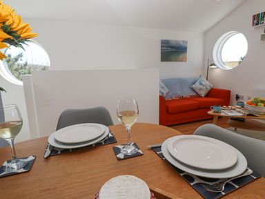 A dining table set with plates and wine glasses in a room with an orange sofa and round windows at Azure Retreat at Una in Carbis Bay near St Ives