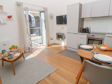 A kitchen and dining area with wooden floor a small dining table set with plates and a wine glass a coffee table with cups and fruit an open door leading outside at Azure Retreat at Una in Carbis Bay near St Ives