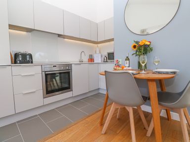 A kitchen with white cabinets oven a small round wooden dining table with two gray chairs and flowers on the table at Azure Retreat at Una in Carbis Bay near St Ives