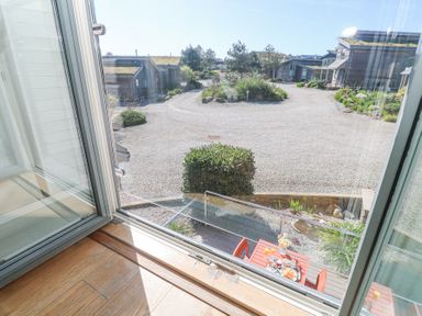 A view through an open window overlooking a gravel driveway with bushes and wooden houses with green roofs and a small table with chairs below Azure Retreat at Una Carbis Bay near St Ives