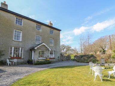 An exterior view of a multi-story house with windows and a small porch surrounded by a gravel driveway and a lawn with outdoor chairs and tables at Cantref House Top Floor in Brecon