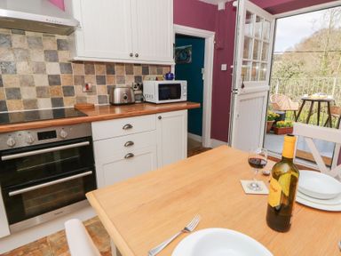 A kitchen with an oven, toaster, microwave, wooden table set with plates, a bottle of wine, and glass with an open door to a balcony with outdoor chairs at Cantref House Top Floor in Brecon