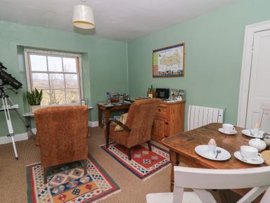 A room with two brown chairs on patterned rugs a wooden table set with cups and eggs a telescope near the window and a wooden cabinet with books and bottles at Cantref House Top Floor in Brecon