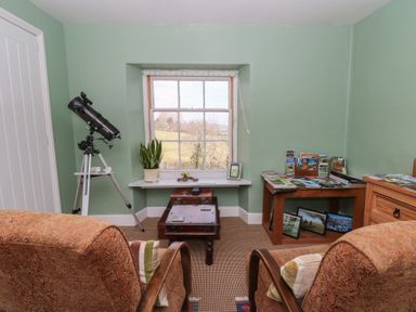 A room with two armchairs facing a window with a telescope on a tripod to the left and a wooden table with brochures to the right at Cantref House Top Floor in Brecon