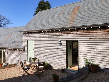 An outdoor patio with gravel floor a small table and two wooden chairs against a wooden paneled building at The Dairy at Wyke Farm Wyke near Lyme Regis