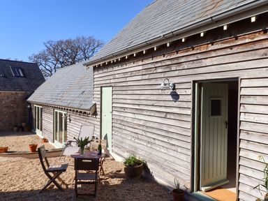 An outdoor gravel area with wooden chairs and a table with a bottle and glasses outside a wooden building at The Dairy at Wyke Farm Wyke near Lyme Regis