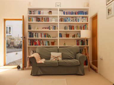 A sitting area with a green sofa and bookshelf filled with books and plants next to an open door and a floor lamp at The Dairy at Wyke Farm in Wyke near Lyme Regis