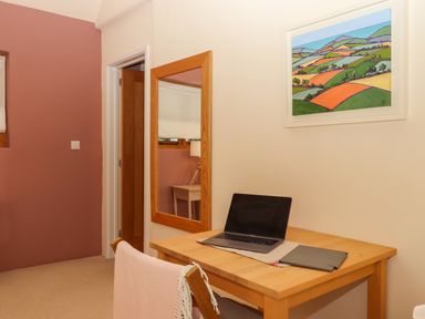 A workspace with a wooden table laptop and tablet against a light wall with a landscape painting and wooden framed mirror at The Dairy at Wyke Farm Wyke near Lyme Regis