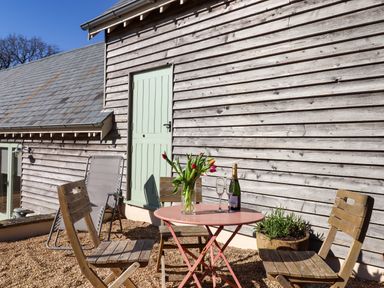 An outdoor patio with a pink round table holding a vase of tulips and a bottle with two glasses surrounded by wooden chairs at The Dairy at Wyke Farm Wyke near Lyme Regis