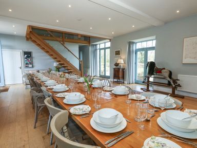 A dining room with a long wooden table set with white dishes and glasses with wooden chairs and a staircase at Stapleford Farm Hooke near Beaminster