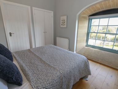 A bedroom with a patterned bedspread two dark pillows a window with stone frame and white doors at Stapleford Farm in Hooke near Beaminster