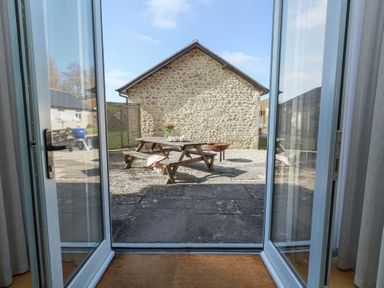View through open glass doors to an outdoor patio area with a wooden picnic table and bench with a vase of flowers on the table at Stapleford Farm in Hooke near Beaminster