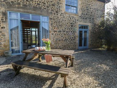 An outdoor seating area with a wooden picnic table and benches in front of a stone building with blue doors and windows at Stapleford Farm in Hooke near Beaminster