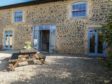 A stone building with blue framed doors and windows and a wooden picnic table with flowers on gravel at Stapleford Farm in Hooke near Beaminster