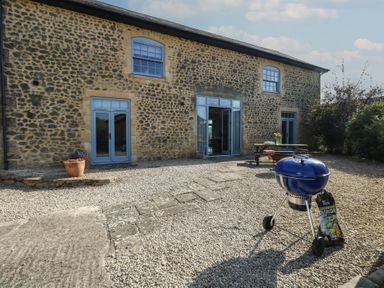 An outdoor patio area with a stone building with blue doors and windows a blue barbecue grill and wooden picnic table with flowers at Stapleford Farm Hooke near Beaminster