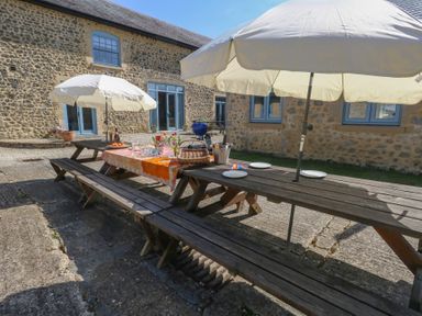 Outdoor patio area with wooden picnic tables umbrellas glasses plates and building with stone walls and blue windows at Stapleford Farm in Hooke near Beaminster