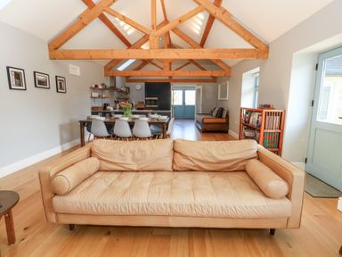 A living room with a tan leather sofa wooden beams dining table and chairs with kitchen and bookshelf at Stapleford Farm in Hooke near Beaminster