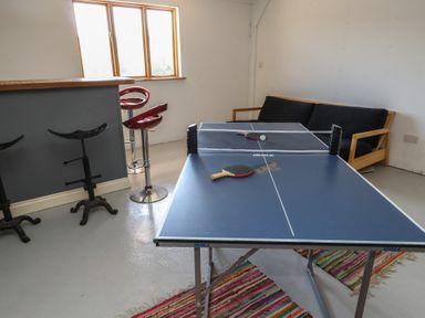 A room with a ping pong table, two paddles, a couch, bar stools, and a window at Stapleford Farm in Hooke near Beaminster