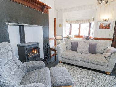 A living room with a wood-burning stove a grey armchair and sofa with cushions in front of a bay window at Bwthyn y Coediwr The Woodcutters Cottage in Tan Y Bwlch Maentwrog