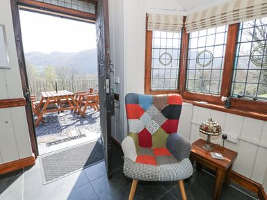 A corner with a colorful patchwork armchair next to a wooden side table with a stained glass lamp by windows and an open door leading to an outdoor wooden table and chairs at Bwthyn y Coediwr (The Woodcutters Cottage) in Tan Y Bwlch Maentwrog