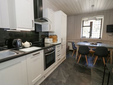 A kitchen area with white cabinets black countertop a stove a kettle and a dining table with black chairs at Bwthyn y Coediwr The Woodcutters Cottage in Tan Y Bwlch Maentwrog