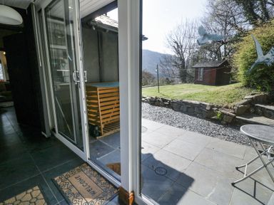 A sliding glass door opens to a stone patio with a metal table and chairs a lawn with a small wooden shed and trees in the background at Bwthyn y Coediwr (The Woodcutters Cottage) in Tan Y Bwlch Maentwrog
