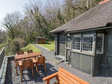 An outdoor area with a wooden table and chairs next to a building with leaded windows and a gravel path leading to a shed at Bwthyn y Coediwr The Woodcutters Cottage in Tan Y Bwlch Maentwrog
