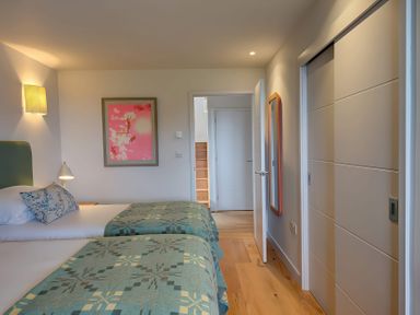 A bedroom with two single beds with patterned green bedspreads a bedside table with a lamp a wall light a framed pink artwork a doorway a wooden floor a full length mirror and a sliding door wardrobe at Villa 4 in Mullion