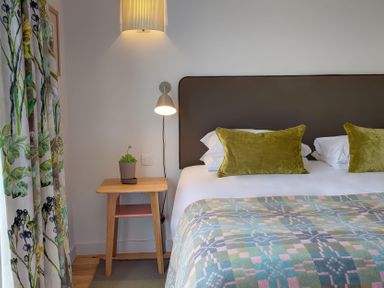 A bedroom with a bed having green pillows and a patterned blanket a wooden bedside table with a plant and two wall lamps at Villa 4 in Mullion
