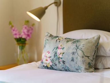 A bed with a floral decorative pillow a wall lamp and flowers in a vase in a bedroom at Villa 5 in Mullion