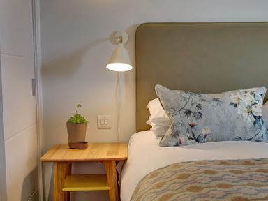 A bedroom corner with a wooden bedside table holding a potted plant a wall lamp and a bed with a floral pillow and patterned blanket at Villa 6 in Mullion