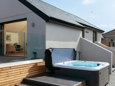 An outdoor space with a hot tub near a wooden deck and a glass railing next to a modern house at Shearwater in Mullion