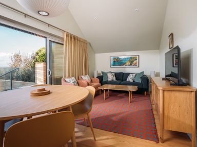 A living room with a wooden dining table and chairs a blue sofa and two armchairs a wooden cabinet with a television and a sliding glass door at Shearwater in Mullion