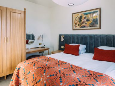 A bedroom with a double bed with orange cushions and blanket a wooden wardrobe and a dressing table with a lamp and round mirror at Shearwater in Mullion