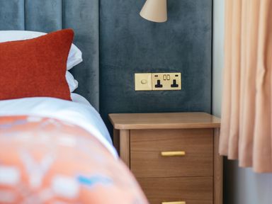 A bedside with a wooden nightstand two drawers orange cushion on bed and electrical outlets on dark blue wall Shearwater Mullion