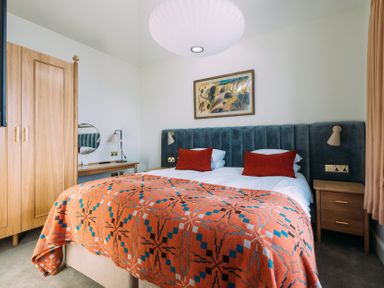 A bedroom with a bed covered by an orange patterned blanket and two red pillows with wooden wardrobe and bedside tables at Shearwater in Mullion