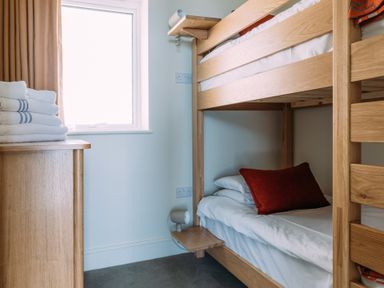 A small bedroom with wooden bunk beds a red pillow folded towels on a wooden chest of drawers and a window at Shearwater in Mullion