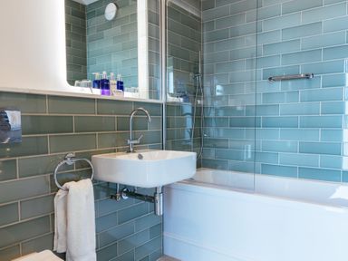 A bathroom with a white sink towel on a ring green tiled walls and a bathtub with a glass shower screen at Shearwater in Mullion