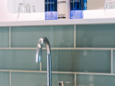A bathroom sink with a chrome faucet green tiles on the wall bottles of Elemis toiletries and upside down glasses on the counter