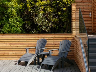 Two gray wooden loungers on a deck with wooden paneling and glass railing surrounded by trees at Shearwater in Mullion
